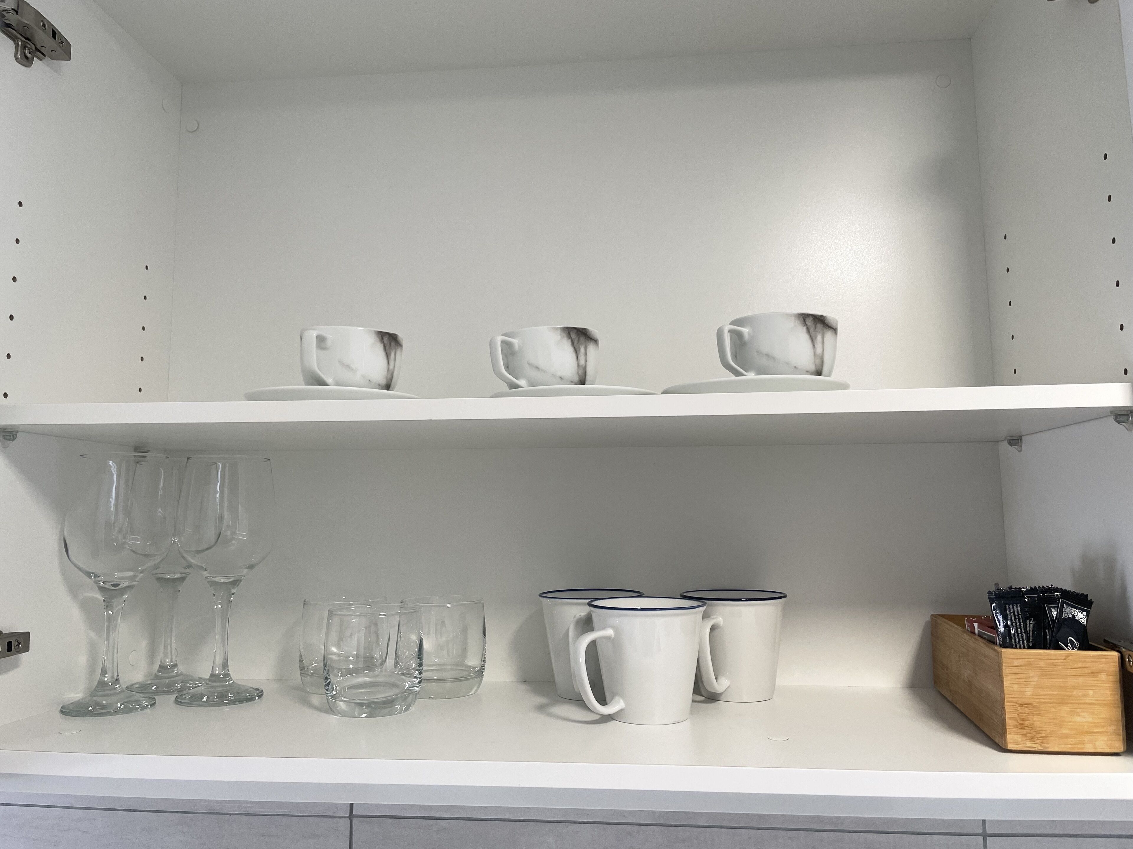 A kitchen shelf with white ceramic cups, clear wine glasses, and a wooden block with black items.