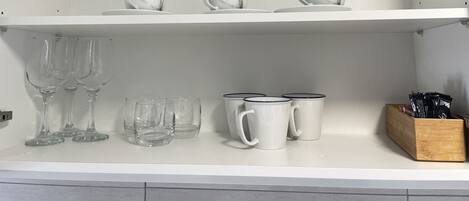 A kitchen shelf with white ceramic cups, clear wine glasses, and a wooden block with black items.