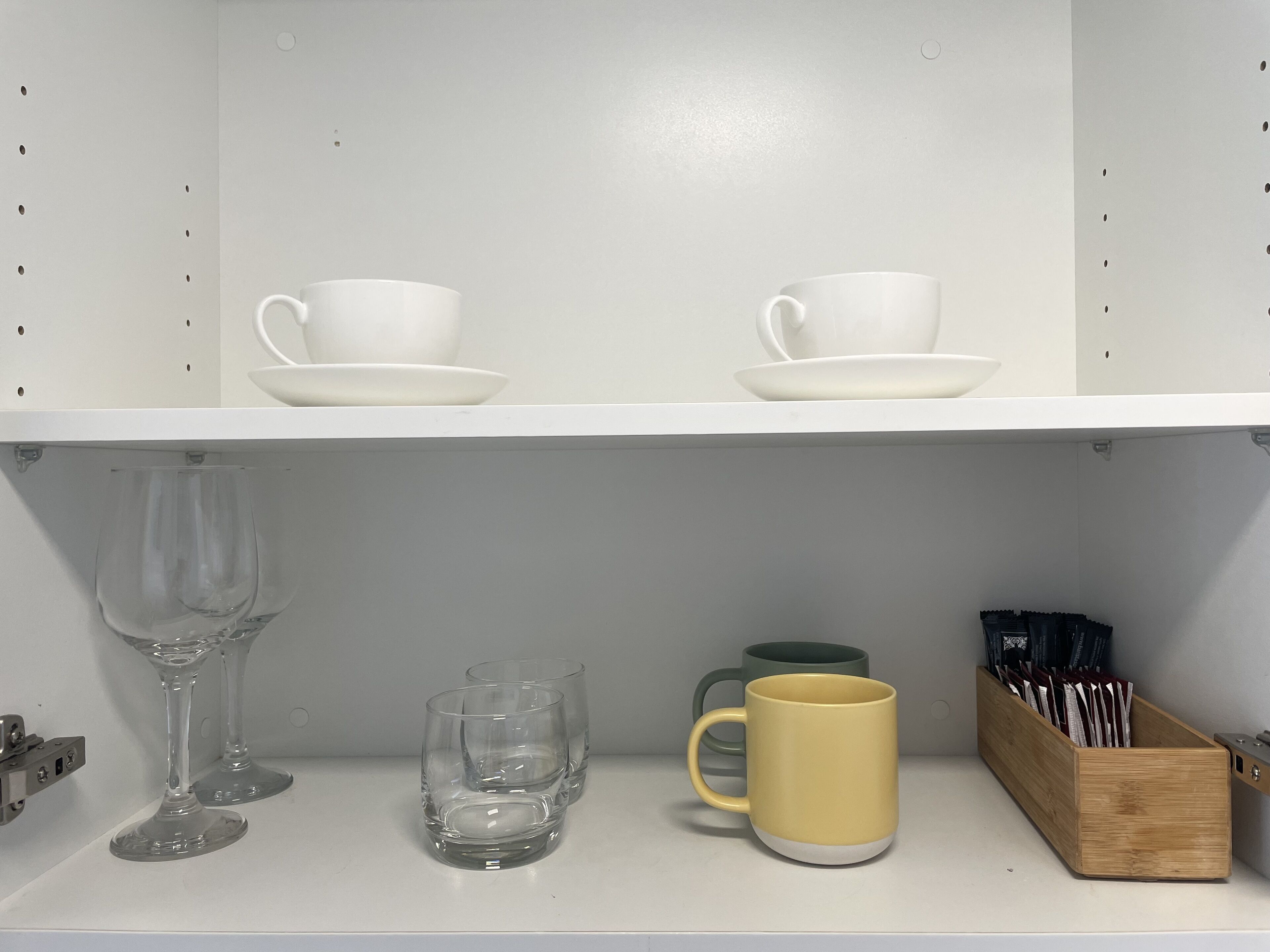 Una estantería con dos tazas y platillos blancos, una copa de vino transparente, un vaso verde y una bandeja de madera con utensilios.