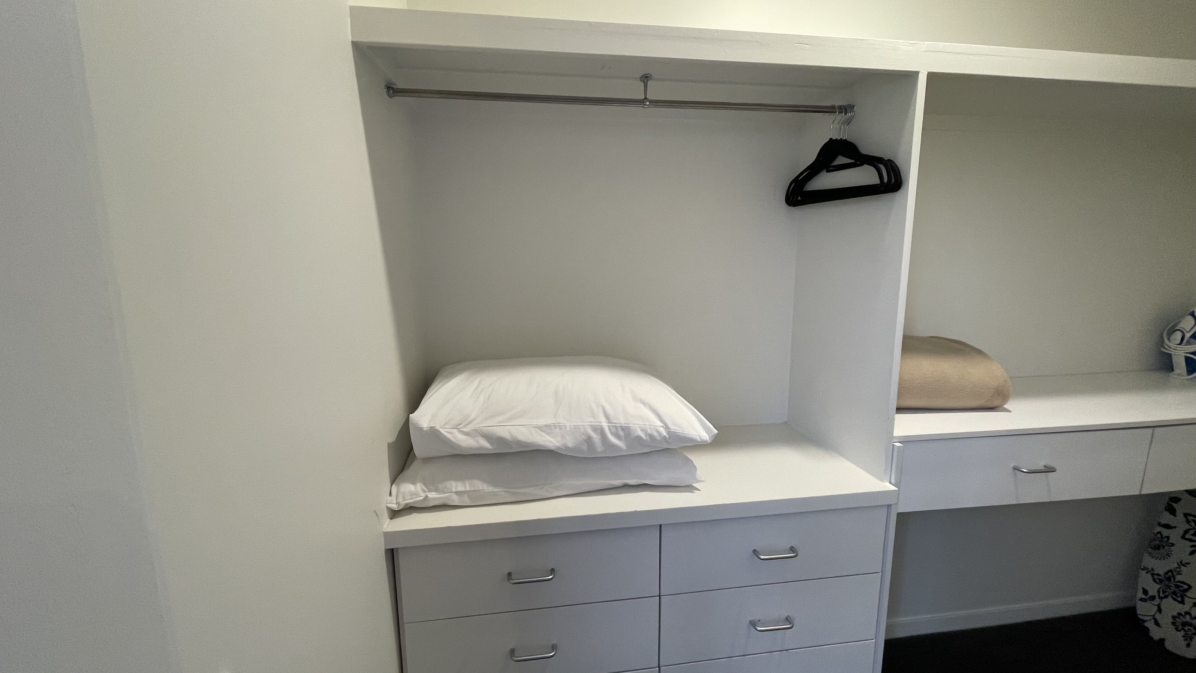 A white walk-in closet with a hanging rod, a bed with white bedding, and a small bedside table with a towel.