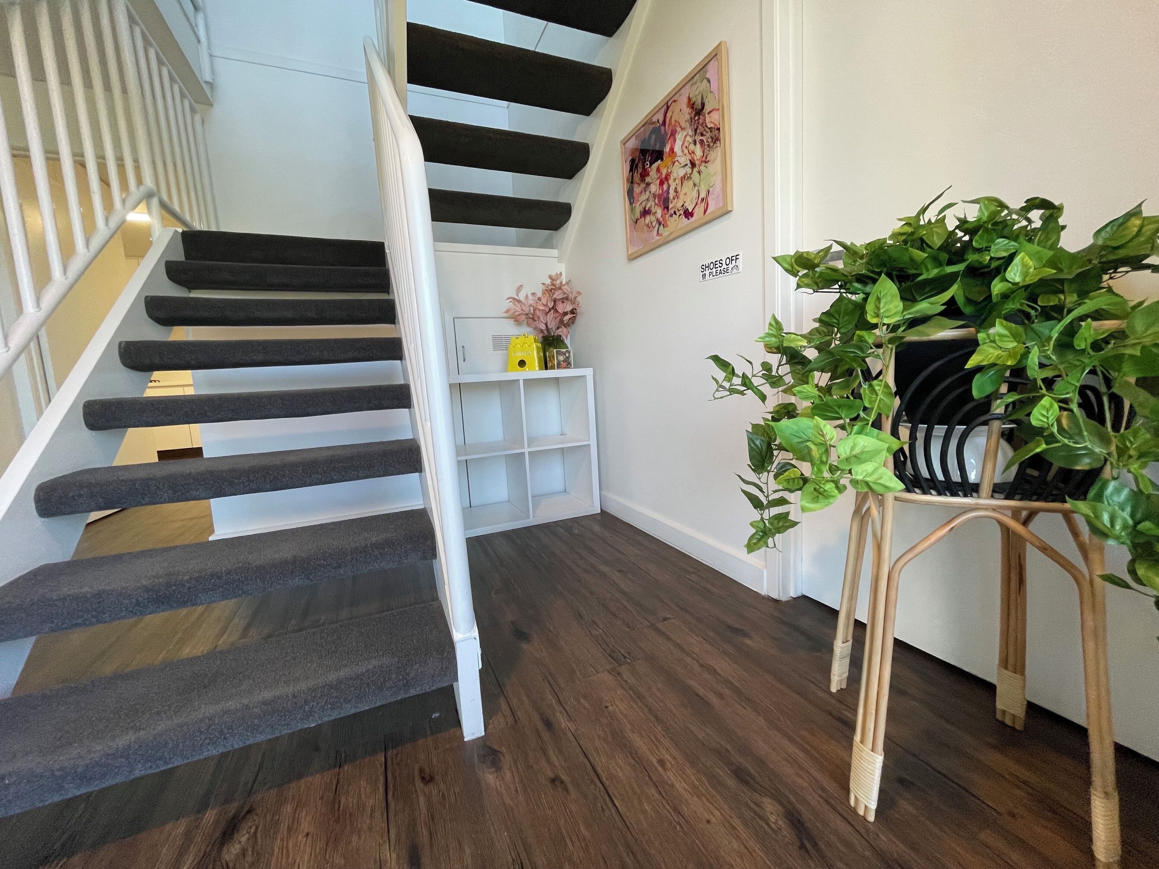 Una escalera con escalones oscuros y barandas blancas, una consola blanca con un jarrón, una planta verde grande y un letrero en la pared que dice 'SHOES OFF'.