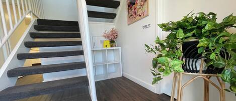 Una escalera con escalones oscuros y barandas blancas, una consola blanca con un jarrón, una planta verde grande y un letrero en la pared que dice 'SHOES OFF'.