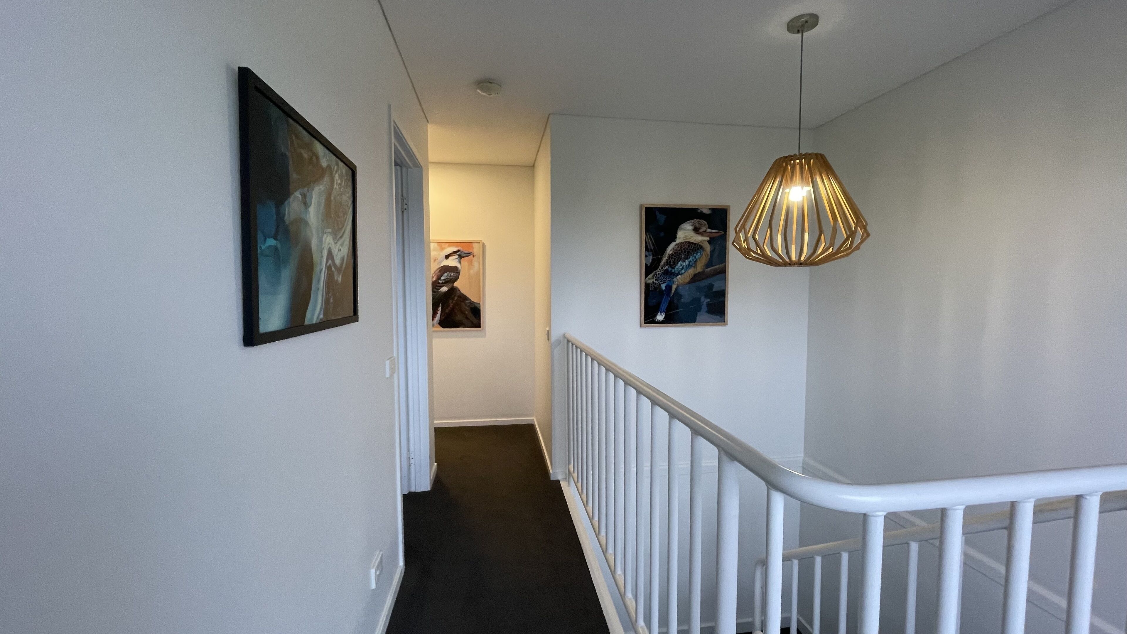 A hallway with a white railing, a hanging pendant light, and framed artwork on the walls.