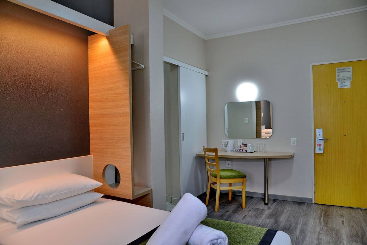 A hotel room with a bed, a desk with a chair, a mirror, and a door.