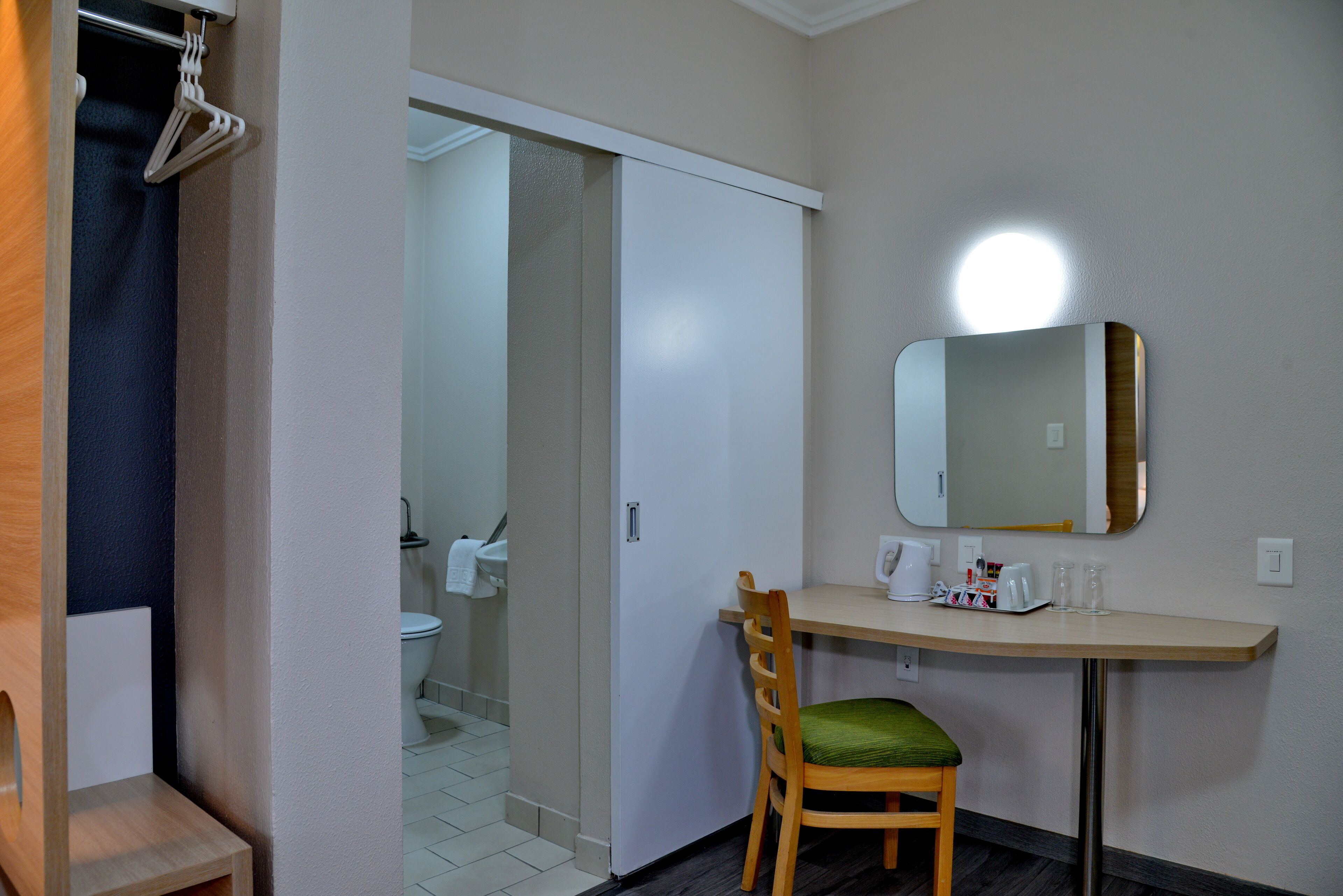 A hotel room with a wooden chair, a small table with a mirror, and a sliding door leading to a bathroom.