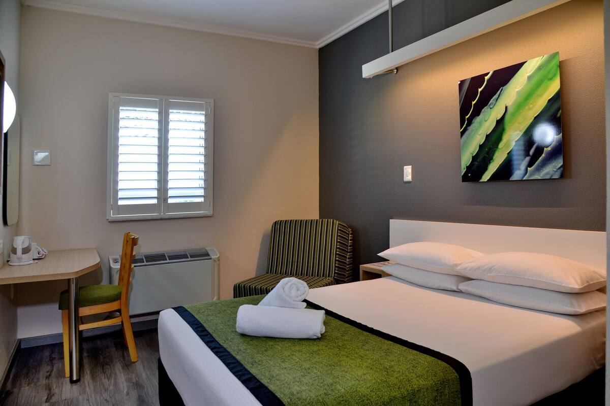 A hotel room with a bed, a desk, a chair, a radiator, and a window.