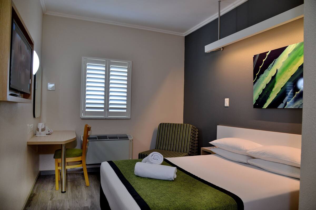 A hotel room with a bed, a desk with a chair, a television, and a window with blinds.