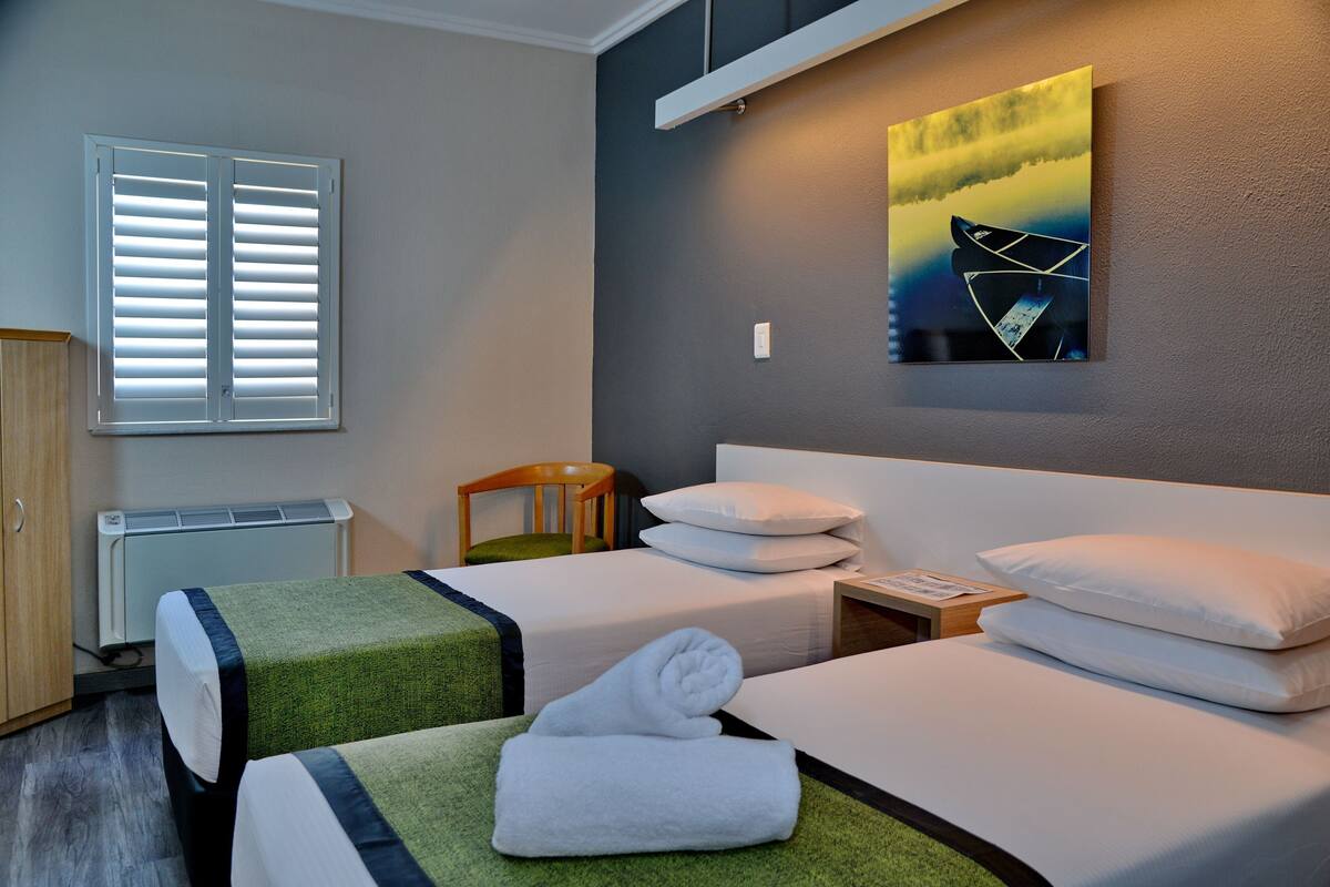 A hotel room with two beds, a chair, a heater, a window with blinds, and a painting on the wall.