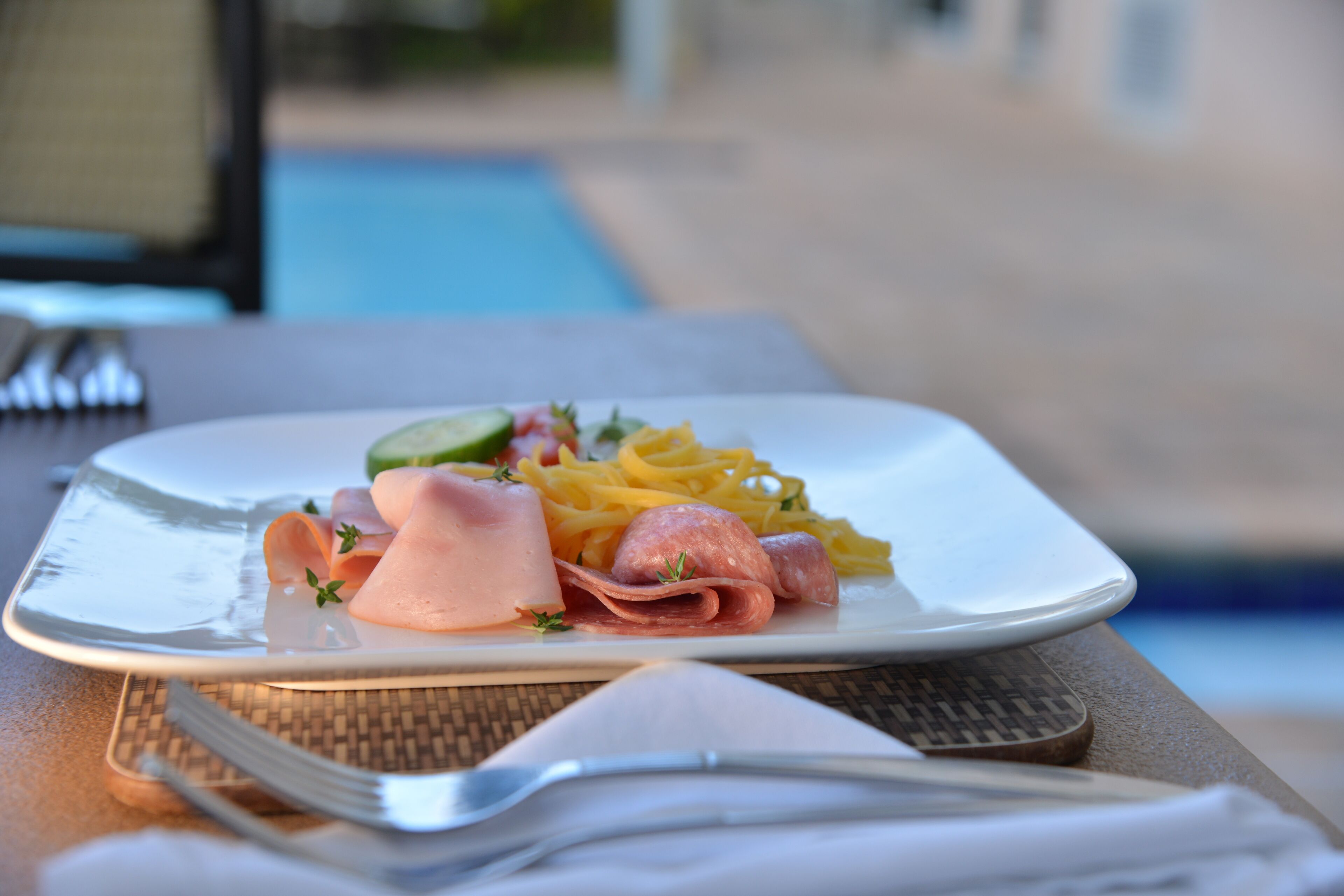 Prato de comida com diversos carnes e vegetais, um garfo e uma faca sobre uma mesa perto de uma piscina.