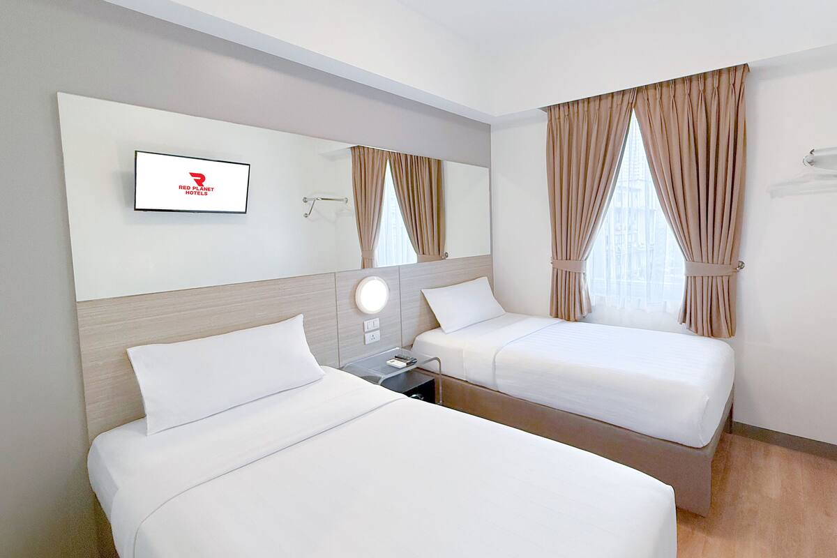 A hotel room with two beds, a TV mounted on the wall, and a window with curtains.
