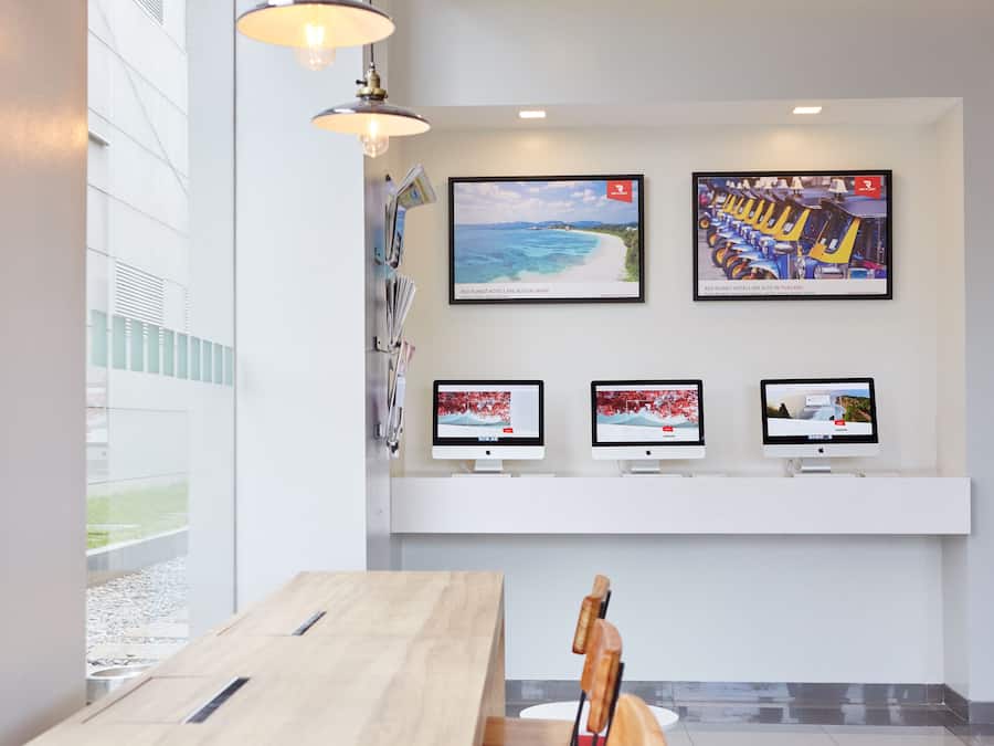 Un vestíbulo de hotel moderno con un mostrador de recepción de madera, tres monitores en una pared y una obra de arte que representa una playa y un tren.