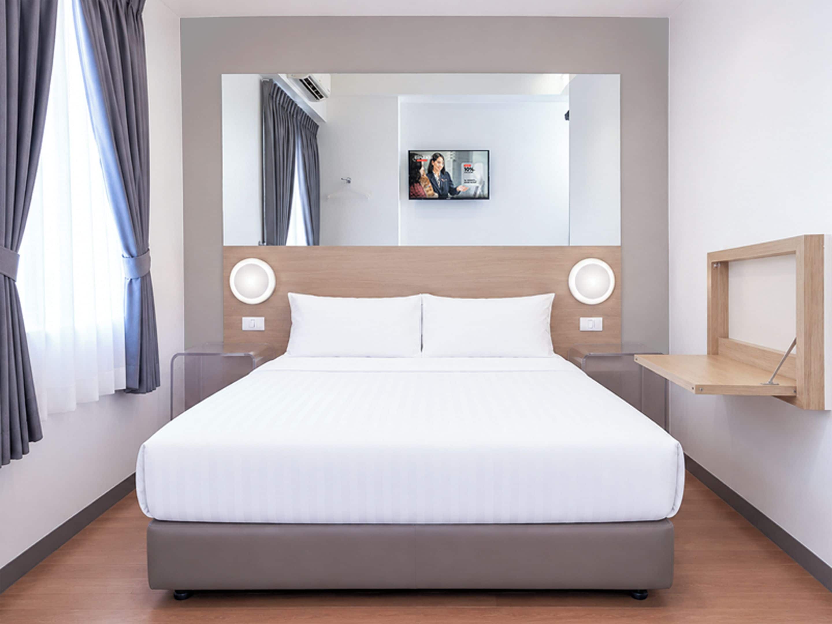 Ein modernes Hotelzimmer mit einem großen Bett, einer Kopfteilbeleuchtung, einem Nachttisch und einem an der Wand befestigten Regal.