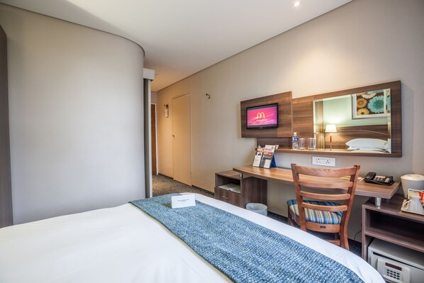 Habitación de hotel con cama, escritorio con computadora, silla, televisor mostrando el logotipo de McDonald's y un espejo.