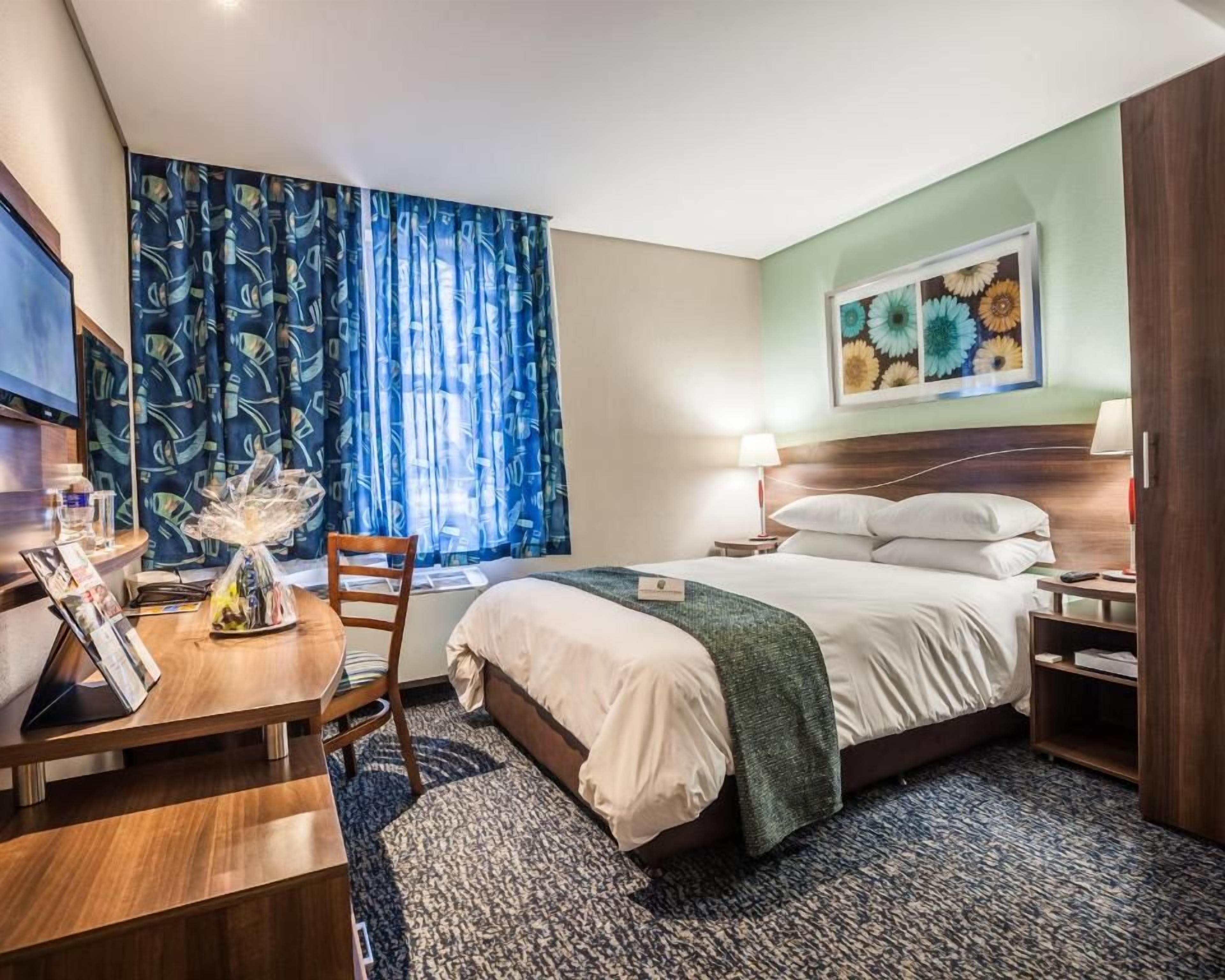 Habitación de hotel con cama, mesita de noche, lámpara y escritorio con silla.