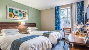 Habitación de hotel con dos camas, un escritorio, una silla, una lámpara, una ventana con cortinas y un cuadro en la pared.