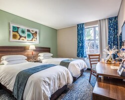 Habitación de hotel con dos camas, un escritorio, una silla, una lámpara, una ventana con cortinas y un cuadro en la pared.