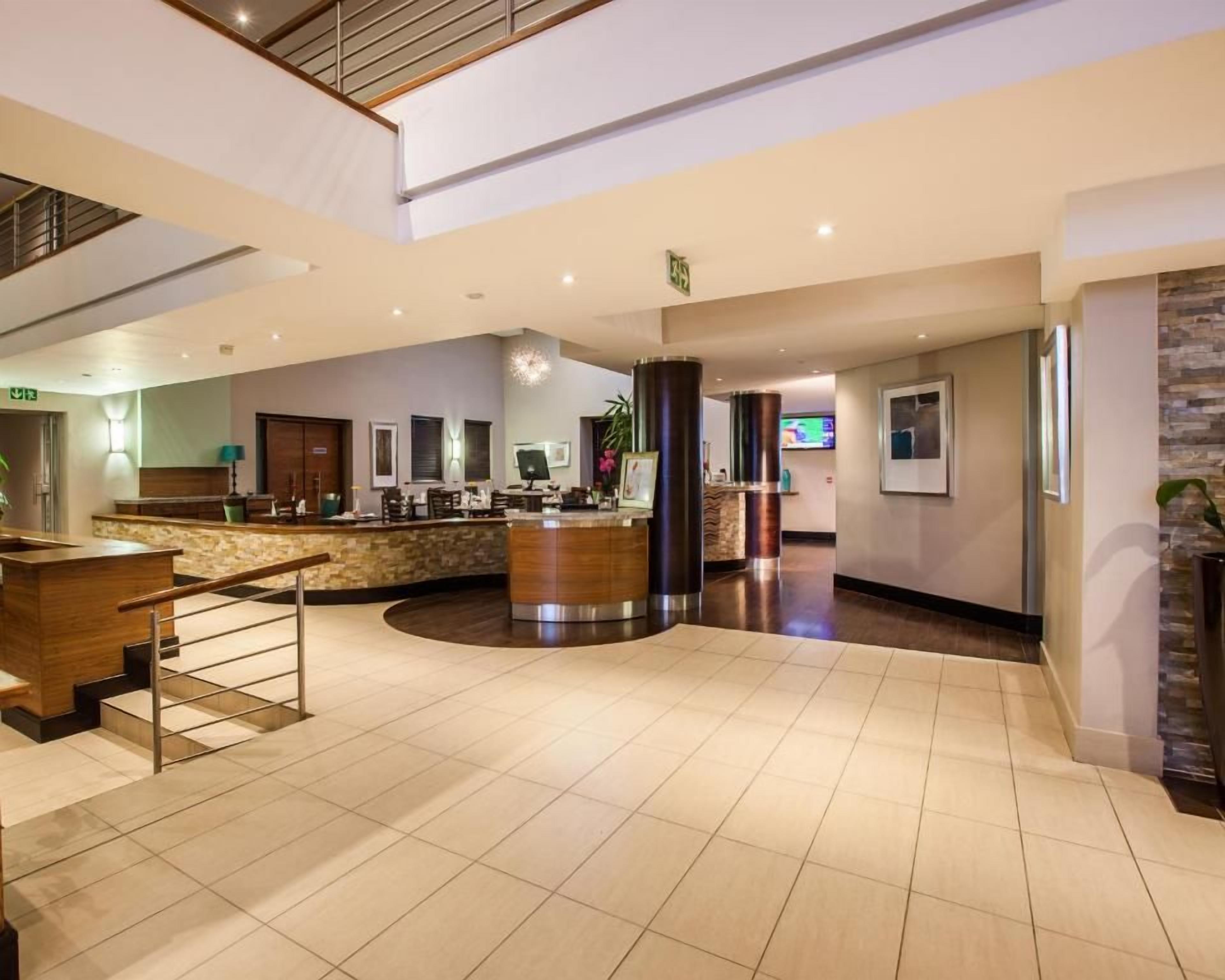 Un vestíbulo de hotel con mostrador de recepción, una escalera y un amplio espacio abierto con techo alto.