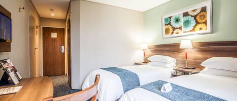 A hotel room with two beds, a wooden desk, a chair, a TV, and a framed floral artwork on the wall.
