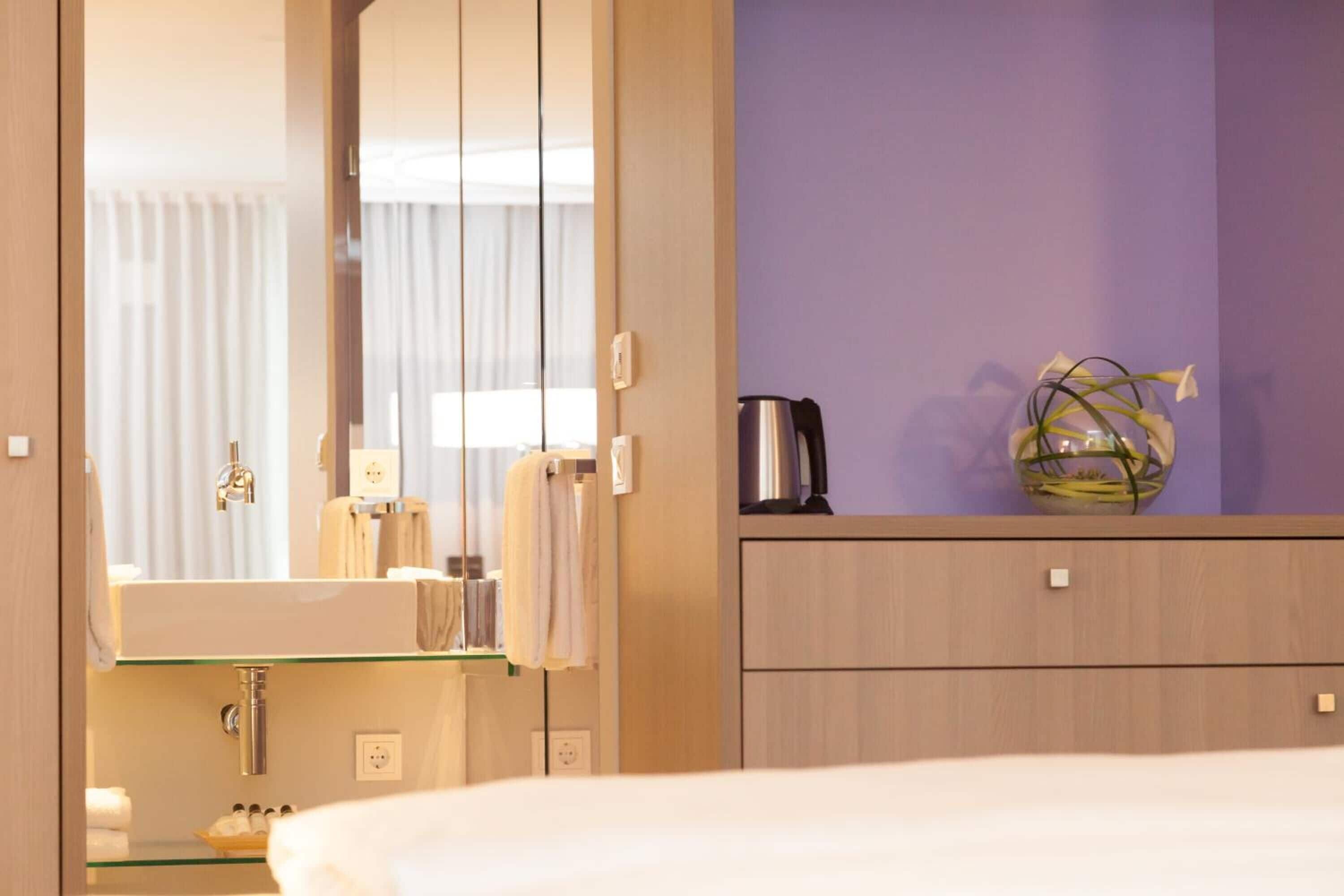 A hotel room with a large mirror, a sink, a coffee maker, and a vase with flowers on the dresser.