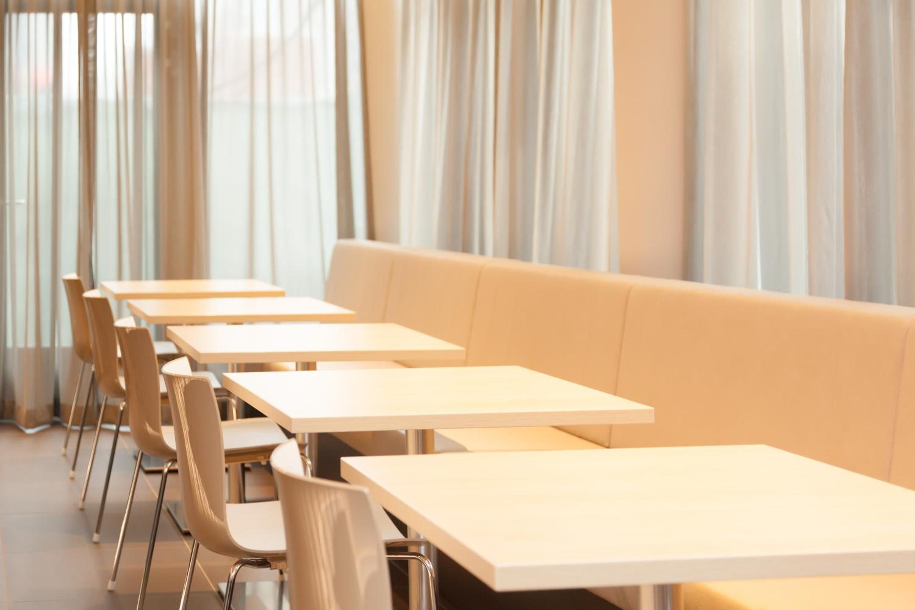 A modern dining area with long tables and chairs, a cushioned bench, and large windows with curtains.