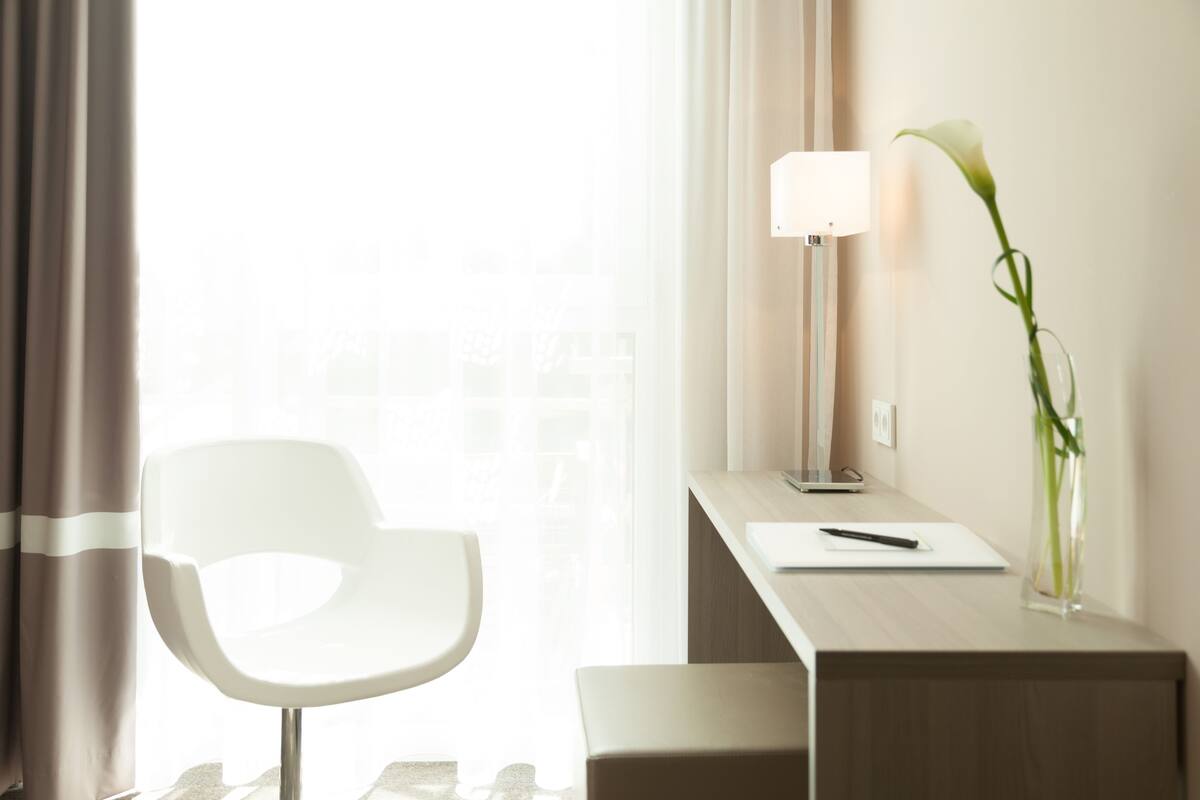 A modern hotel room with a white chair, a desk with a lamp, and a vase with a plant.