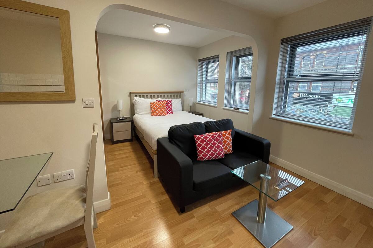 Ein kompaktes Hotelzimmer mit Bett, kleinem Schreibtisch, einer Couch und einem Glastisch. Zwei Fenster mit Jalousien und eine Deckenleuchte sind vorhanden.
