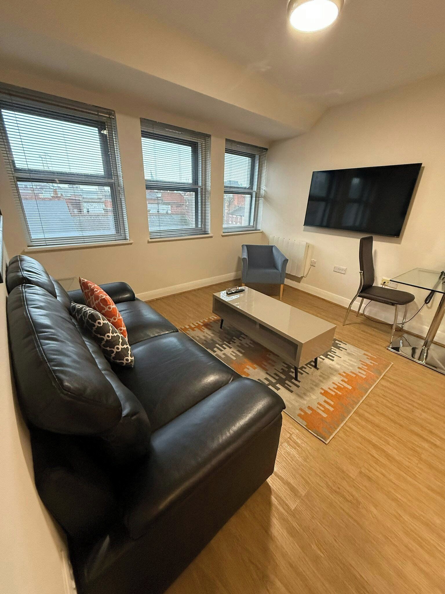 A modern living room with a black leather sofa, a glass coffee table, and a grey armchair. There are three windows with blinds, a flat-screen TV mounted on the wall, and a radiator.