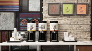 Un espace dédié au café, équipé de trois distributeurs de café étiquetés « Café léger », « Café décaféiné » et « Eau chaude ». Des œuvres d’art encadrées ornent le mur derrière cet espace.