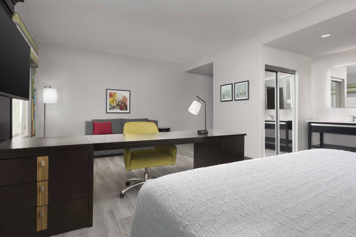 Una habitación de hotel moderna con una cama grande, un escritorio con una silla, una lámpara de escritorio, un sofá y un espejo amplio.