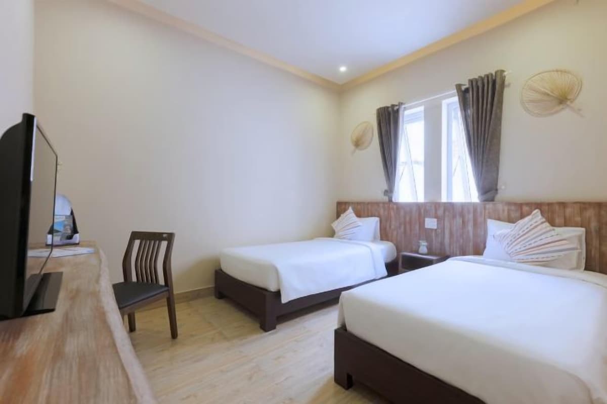 Habitación de hotel con dos camas individuales, un escritorio con silla, televisión y ventana con cortinas.