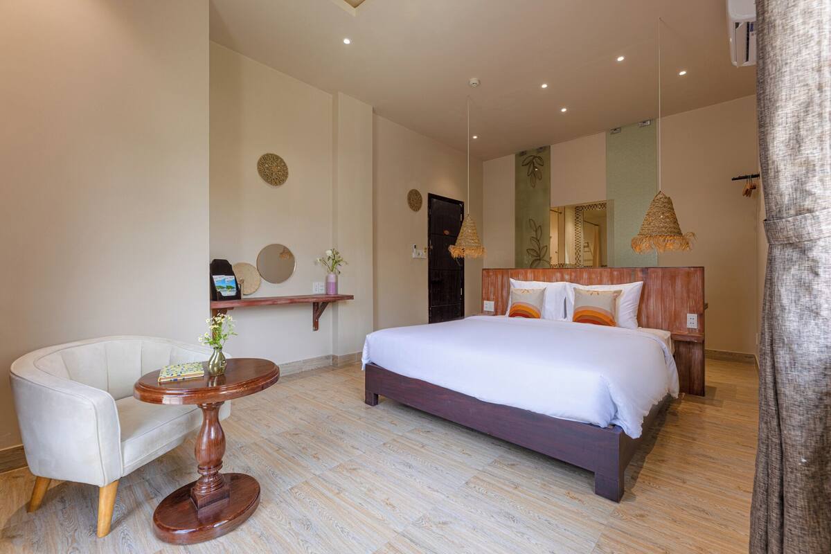 Un dormitorio con cama, una silla, una mesa redonda de madera y una lámpara colgante.