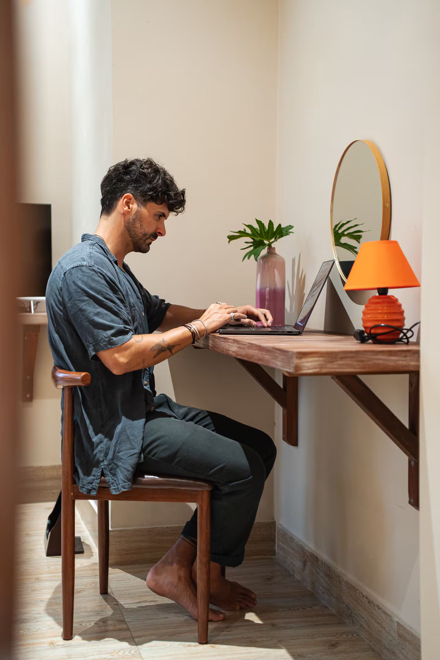 A person seated at a wooden desk with a laptop, a vase with a plant, and a lamp.