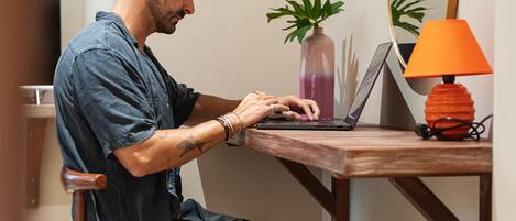 A person seated at a wooden desk with a laptop, a vase with a plant, and a lamp.