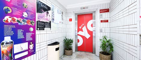 Un couloir aux murs recouverts de carreaux blancs, une porte d’ascenseur rouge et une poubelle. Des affiches sont accrochées au mur, et des plantes en pot y sont disposées.