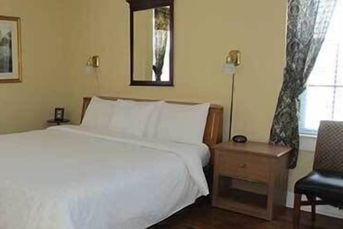 A bedroom with a large bed, a nightstand, a chair, a mirror, and a framed picture on the wall.