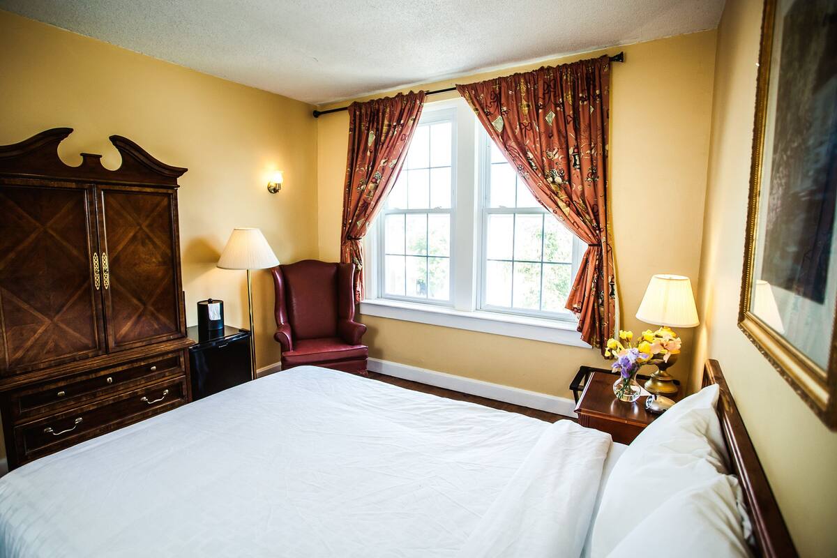 A hotel room with a large bed, a wooden wardrobe, a red armchair, a window with curtains, and a nightstand with a lamp and flowers.