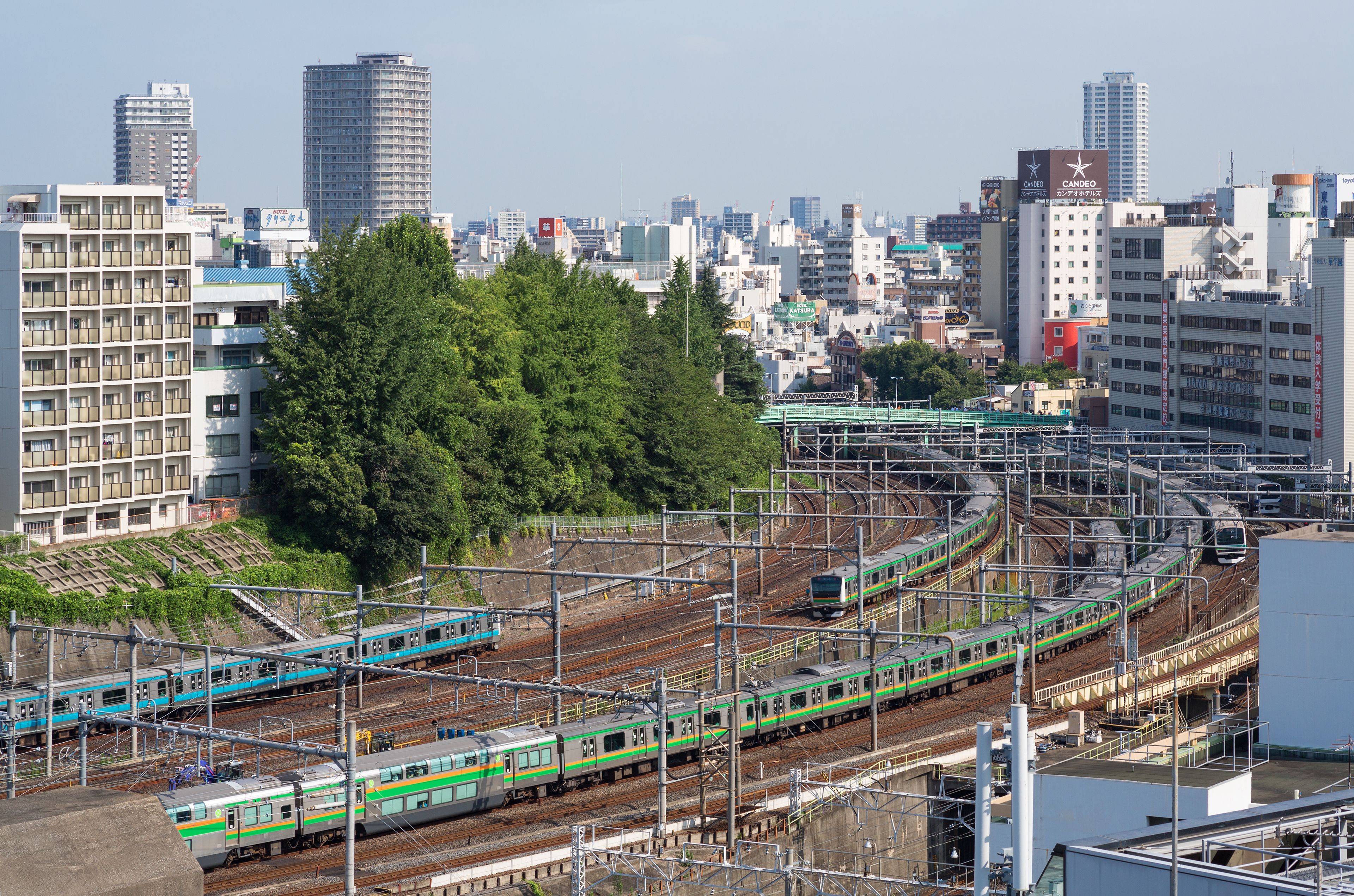 A train station with multiple tracks and trains, surrounded by urban buildings and greenery.