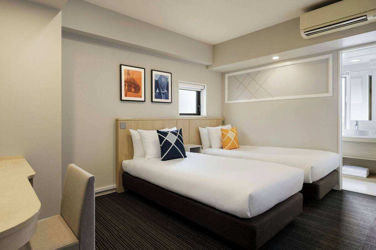 Quarto de hotel com duas camas, uma escrivaninha, uma cadeira e uma janela.