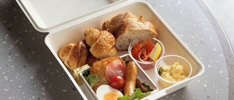 A meal in a white takeout container with croissants, a boiled egg, a tomato, a piece of ham, and a small container of sauce.