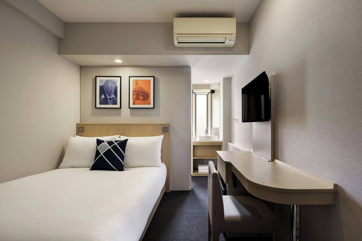 Quarto de hotel com uma cama, uma escrivaninha, uma televisão e duas molduras na parede.