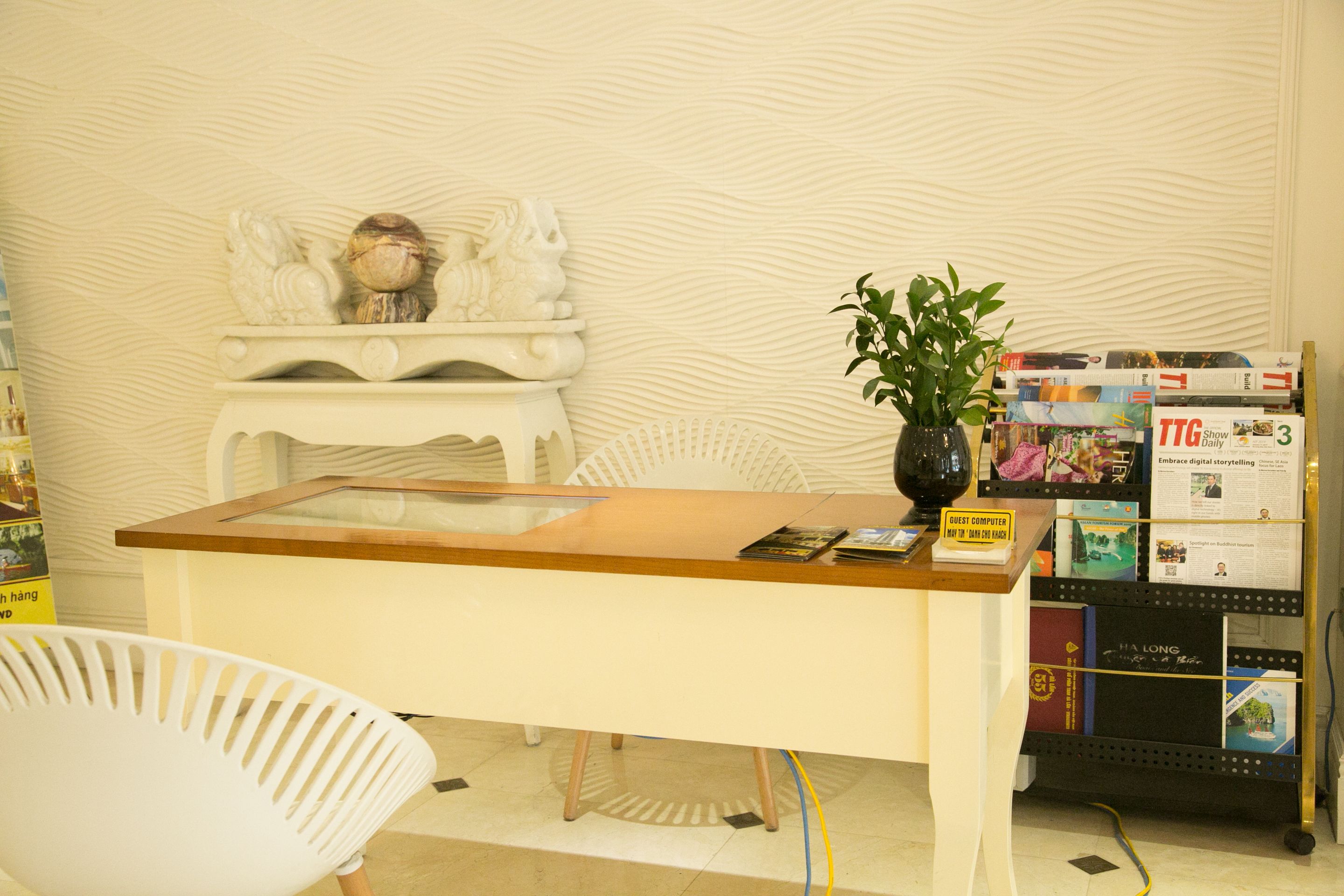 Una reception con un bancone in legno, una sedia bianca, un piccolo tavolino con un oggetto decorativo e una libreria con riviste e libri.