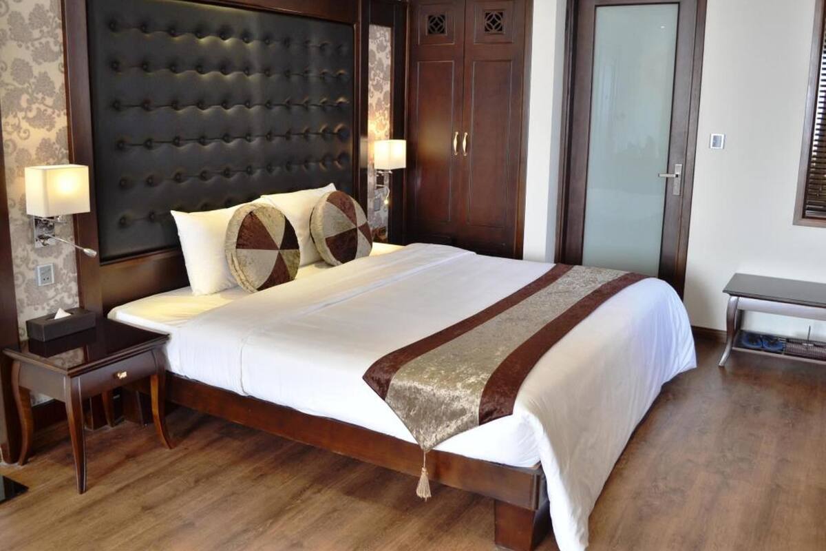 A hotel room with a large bed, a wooden headboard, two bedside tables with lamps, a wardrobe, and a door.