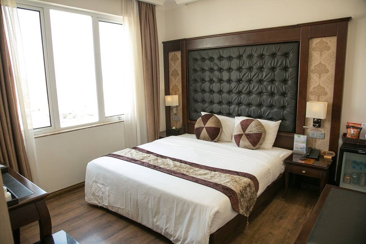 A hotel room with a large bed, a nightstand with a lamp, a small table with a phone and a menu, and a minibar.