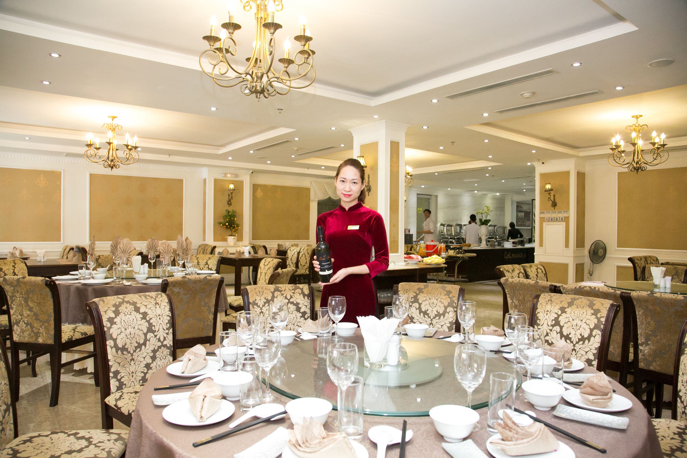 A well-lit dining area with round tables set for a meal, a woman in a red dress standing, and chandeliers overhead.