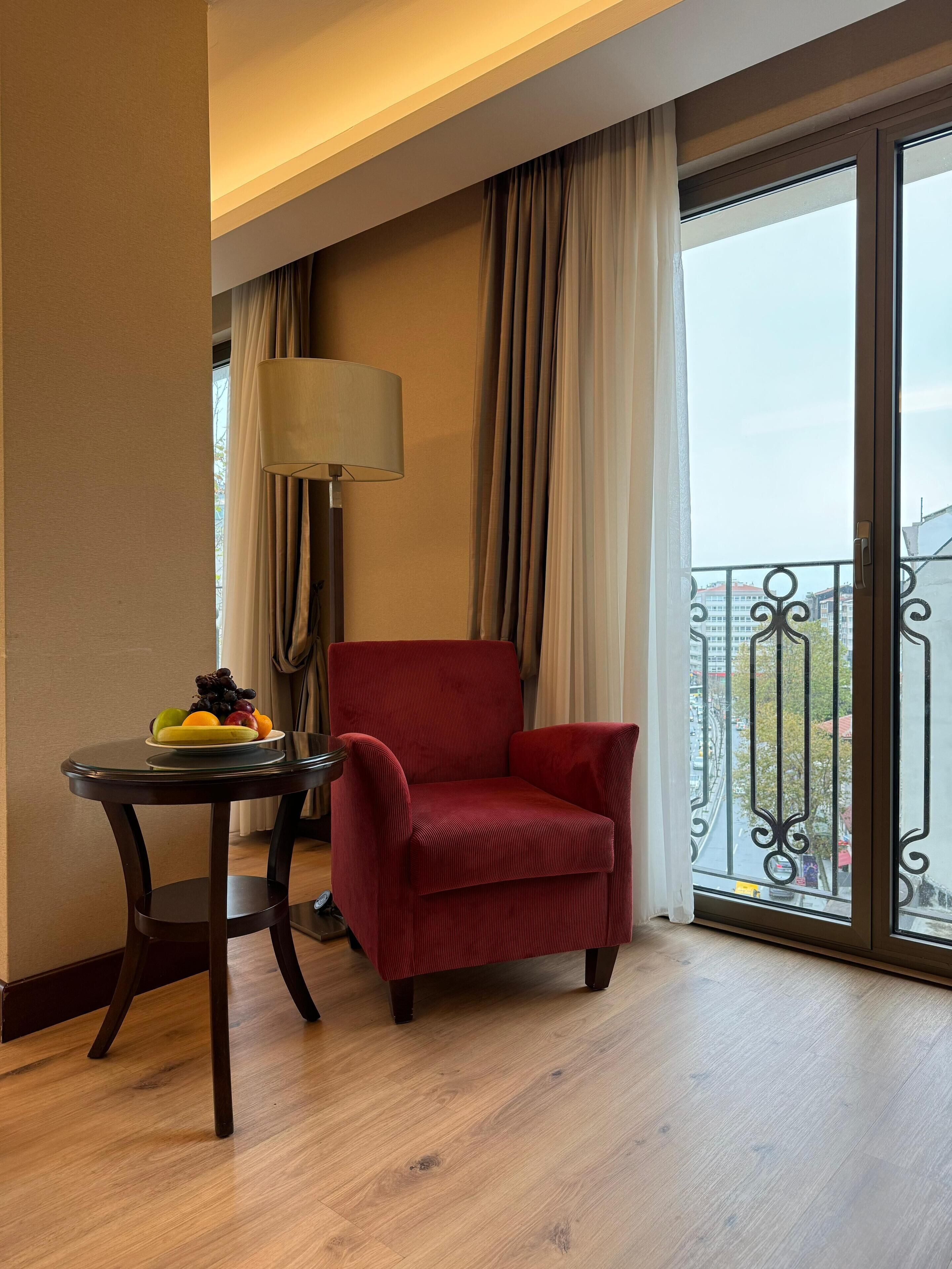 A hotel room with a red armchair, a small round table with fruit, a lamp, and a view of the city through a balcony with a wrought iron railing.