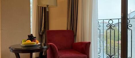 A hotel room with a red armchair, a small round table with fruit, a lamp, and a view of the city through a balcony with a wrought iron railing.