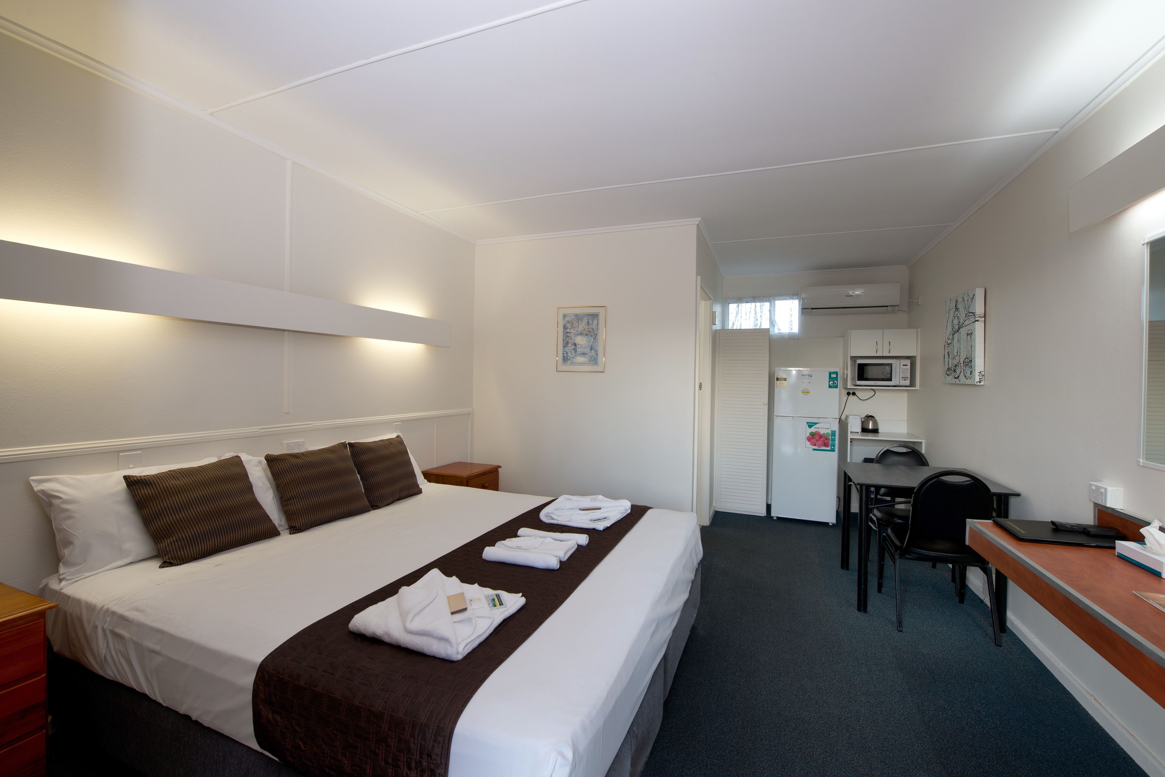 A hotel room with a neatly made bed, a desk with a chair, a microwave, and a refrigerator.