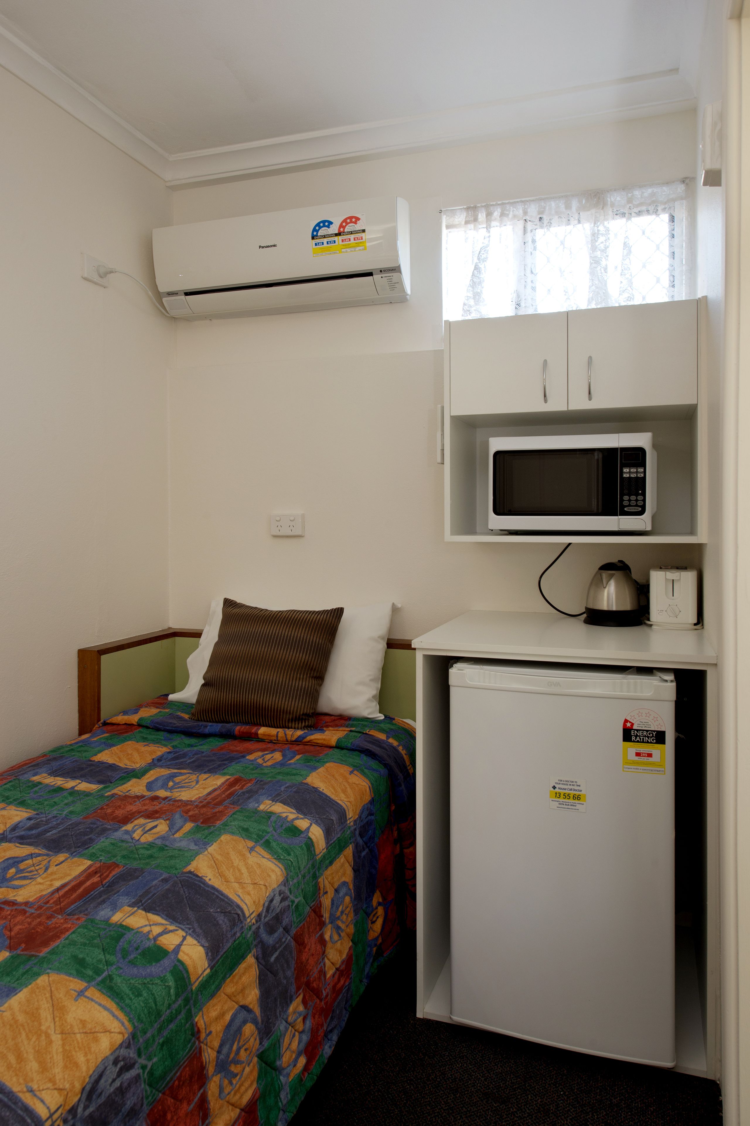 A small, compact room with a bed, a microwave, a refrigerator, and an air conditioning unit.