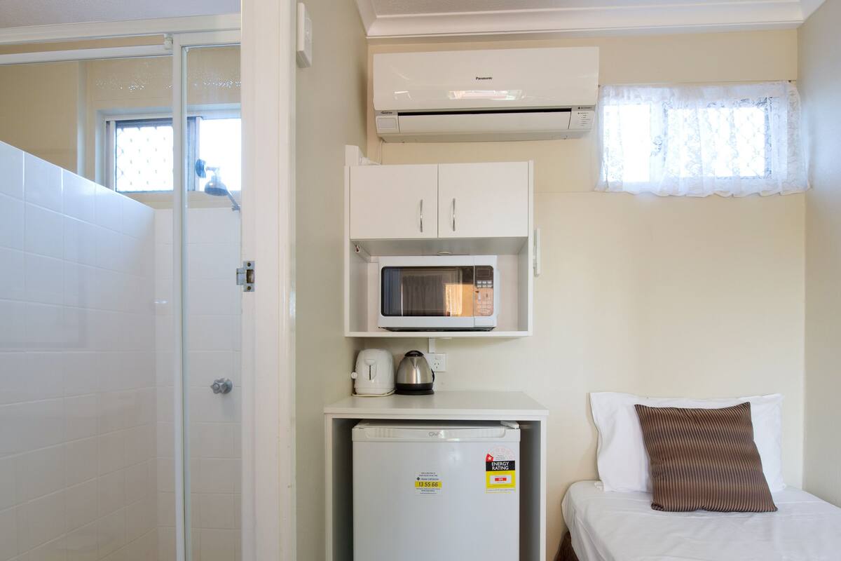Quarto de hotel pequeno com chuveiro, um kitchenette, uma cama e uma janela com cortinas.