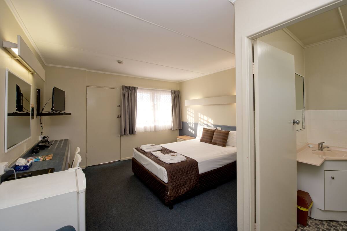 Quarto de hotel com cama, secretária com computador, espelho, banheiro com pia e janela com cortinas.